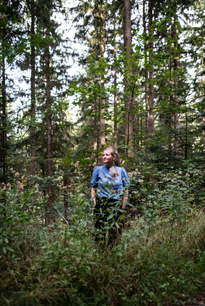 Frau (Katrin Aufenanger) steht im Wald, in einer blauen Bluse, umgeben von Pflanzen und Bäumen, neue Perspektiven und Blickwinkel entdecken
